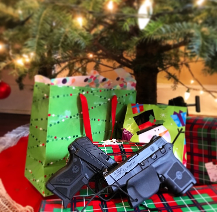 Two pistols outfitted with Clipdraw trigger guards and belt clips lying on a tree skirt surrounded by presents under a lit Christmas tree.